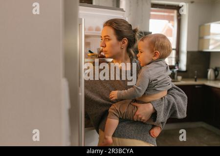 Mother carrying baby boy looking into refrigerator in kitchen at home Stock Photo