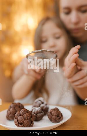 Mother assisting daughter sifting powdered sugar on chocolate pine cones Stock Photo
