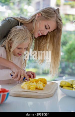 Smiling woman assisting daughter cutting pineapple on cutting board at home Stock Photo