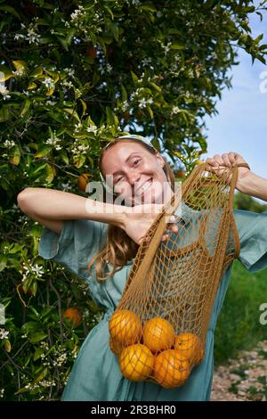 Cheerful woman with fresh oranges in mesh bag at orchard Stock Photo
