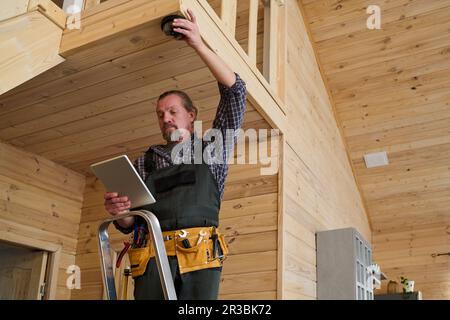 Mature adjuster or fitter in workwear looking through data on tablet screen while standing on step ladder and installing alarm system Stock Photo