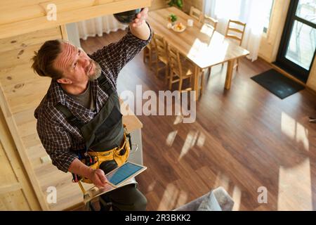 Mature fitterman in coveralls and checkered shirt fixing or adjusting alarm bell and using tablet with settings data on screen during work Stock Photo