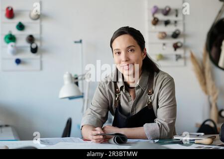Young smiling brunette woman in workwear sitting by table and looking at camera while working with textile or leather samples and sketches Stock Photo