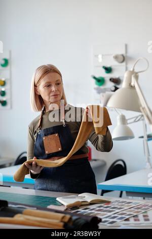 Blond mature craftswoman or designer in apron choosing textile for sewing new trendy apparel while working over seasonal fashion collection Stock Photo