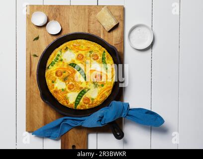 Overhead View of Egg Frittata with Tomato, Peas and Cheese In A Cast Iron Pan on Wood Cutting Board Stock Photo