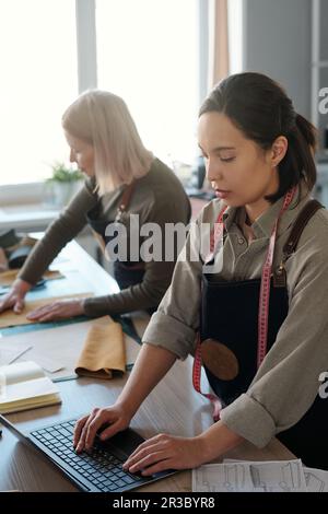 Focus on young brunette female leatherworker typing on laptop keyboard while checking online orders of clients against mature colleague Stock Photo