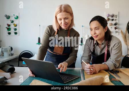 Smiling mature craftswoman making presentation of new fashion trends to young colleague during discussion of creative ideas at meeting Stock Photo