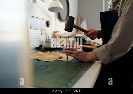 Professional female tanner keeping hammer over small steel shaft while pressing it down leather piece during work in craftshop or studio Stock Photo