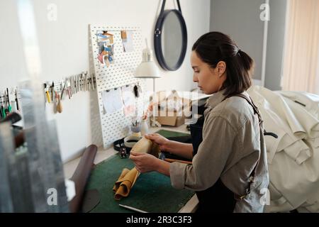 Young brunette woman standing by workplace in studio or craftshop and stretching edges of suede or leather while checking its durability Stock Photo