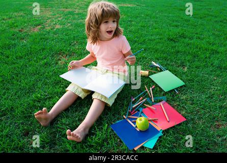 Kids Exam Study. Funny little student boy reading book outdoors, lying on lawn in park, studying at School backyard. Stock Photo