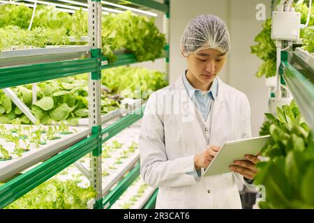 Young serious Asian scientist in lab coat scrolling through online information in tablet while standing between vertical trusses Stock Photo