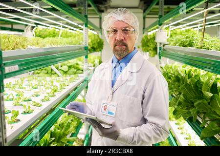 Mature professional biologist in lab coat, protective cap, gloves and eyeglasses looking at camera while using tablet in greenhouse Stock Photo