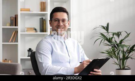 Happy professional psychologist man sitting in chair writing on clipboard, looking and smiling at camera while working in modern office Stock Photo