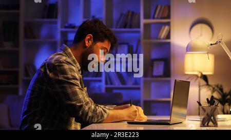 A young Arab student works on his laptop in the evening in the living room. A man with a beard works in IT from home, taps on the keyboard, writes in Stock Photo