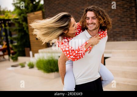Handsome long hair man carrying the pretty young woman on his back in front of brick house Stock Photo