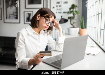 Disappointed young businesswoman working in cozy office or cafe, holding credit card and trying to make purchase Stock Photo