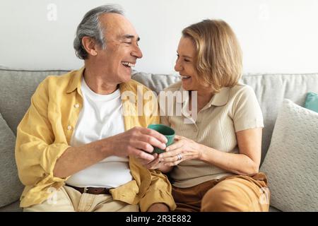 Cheerful senior spouses drinking coffee sitting on sofa, talking and laughing at home, enjoying conversation and hot tea Stock Photo
