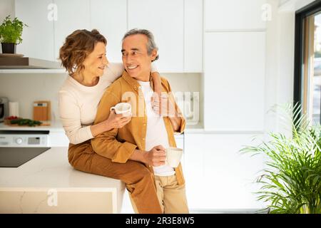 Loving senior married couple enjoying morning coffee at home, happy woman sitting on kitchen table, hugging her husband Stock Photo