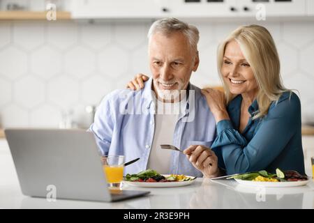 Smiling Elderly Spouses Relaxing With Laptop While Having Lunch In Kitchen Stock Photo