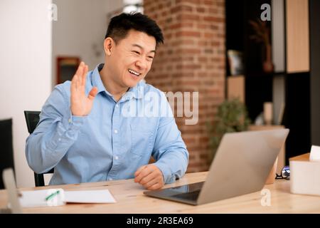 Smiling asian middle aged businessman waving hand at laptop webcamera, having online meeting at home Stock Photo