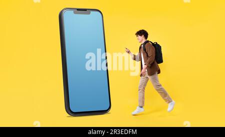 Young Man Walking To Large Smartphone Using Phone, Yellow Background Stock Photo