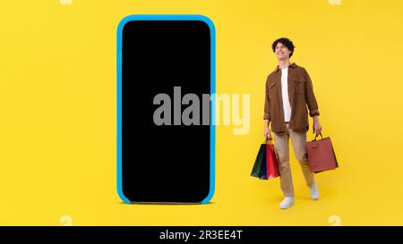 Man With Shopper Bags Standing Near Big Smartphone, Yellow Background Stock Photo