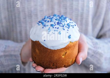 Hands hold Easter cake with white topping and blue sprinkles. Woman holding traditional Russian Easter cake. Homemade pie for sp Stock Photo