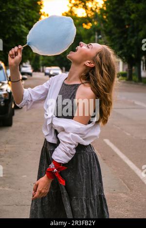 Cute and pretty young girl or student, eats and poses with sugar blue candy cotton on town street, concept happy times. Lifestyl Stock Photo
