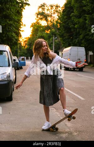 Millennial Woman in dress riding a skateboard on street. Skater girl on a longboard. Cool female skateboarder at sunset. Carefre Stock Photo