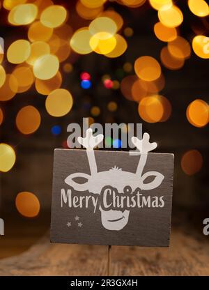 A wooden sign with a reindeer face and the words Merry Christmas against a blurry light background, seasonal holiday concept, Christmas card Stock Photo