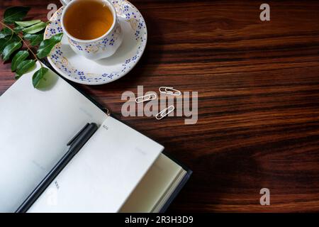 Flat lay design of work desk with teacup, notebook, pen, paper clips, and leaves on wooden background. Stock Photo