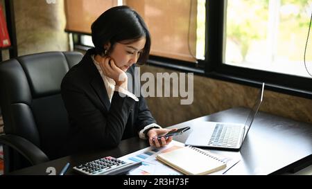 A focused Asian businesswoman hand on chin, reading an online article on the website through her smartphone at her desk. Stock Photo