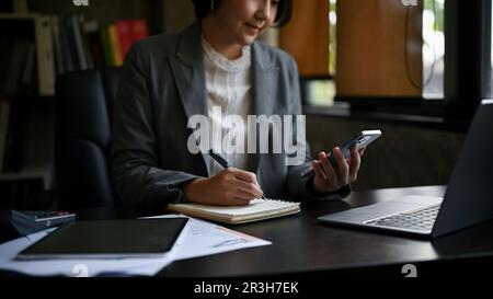 Cropped image of a professional millennial Asian businesswoman or female boss searching informations on internet via her phone and taking notes on her Stock Photo