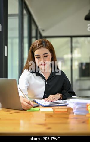 Portrait of a smiling and beautiful millennial Asian businesswoman working on business documents and analyzing financial reports at her desk. Stock Photo