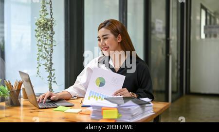 Smiling and happy millennial Asian businesswoman or female financial analyst working on her financial work on laptop and checking reports at her desk. Stock Photo