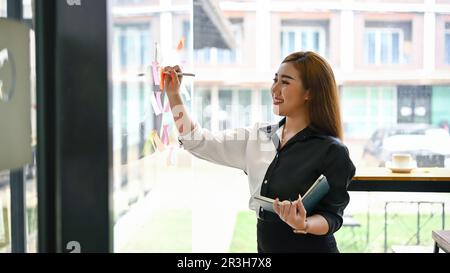 A creative millennial Asian female marketing assistant writing her creative idea on a sticky note on the glass wall. Stock Photo