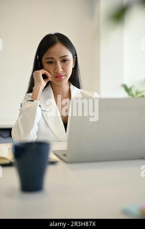 Concentrated and professional Asian businesswoman or female manager reading project details on her laptop computer, working at her desk. Stock Photo
