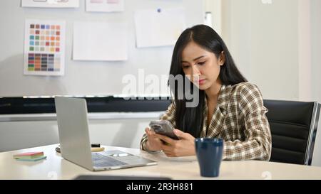 Attractive young Asian female designer checking messages on her smartphone at her desk in the office. Stock Photo