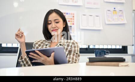 Attractive young Asian female web designer using her digital tablet at her desk in the office. UX UI developer concept Stock Photo