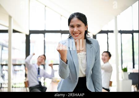 Excited and cheerful young Asian businesswoman smiling and showing clenched fist, rejoicing and celebrating her career success. Stock Photo