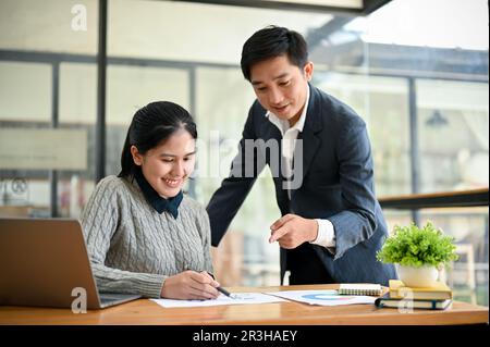 A kind and professional millennial Asian male boss is training and briefing a new project to a young female employee in the office. Stock Photo