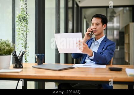 A focused and professional millennial Asian businessman looking some details on a paper while talking on the phone with his business partner. Stock Photo