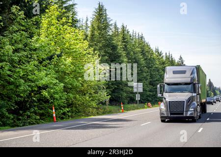 Industrial grade powerful pro big rig dark gray semi truck tractor transporting loaded long container on semi trailer driving on the straight highway Stock Photo