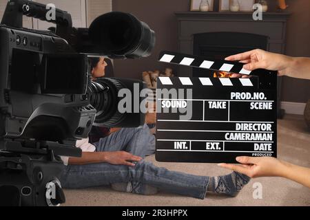 Shooting movie. Second assistant camera holding clapperboard near video camera in front of couple (actors) in room with fireplace (film set) Stock Photo