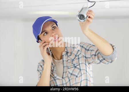 female technician inspecting surveillance camera and talking on smartphone Stock Photo