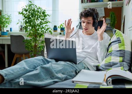 Young guy in headphones using laptop for video communication, lying at home Stock Photo