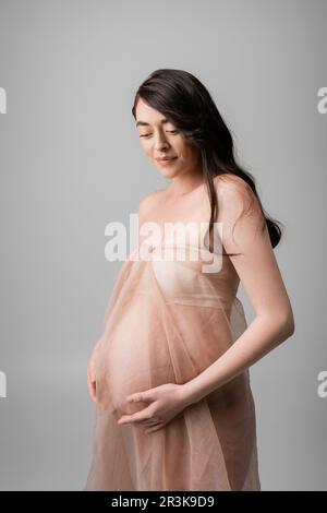 charming brunette mother-to-be in airy chiffon cloth and golden bracelets touching belly and smiling isolated on grey background, maternity fashion co Stock Photo