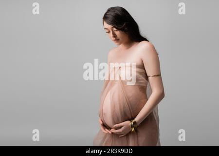 elegant expecting mother with wavy brunette hair, standing in beige chiffon cloth and golden bracelets isolated on grey background, maternity fashion Stock Photo