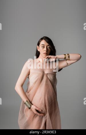 pretty and romantic mother-to-be looking at camera while posing in golden bracelets and airy chiffon draping isolated on grey background, maternity fa Stock Photo