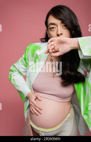 brunette pregnant woman in crop top, fashionable blazer and beads belt holding hand near face, touching tummy and looking at camera isolated on pink b Stock Photo